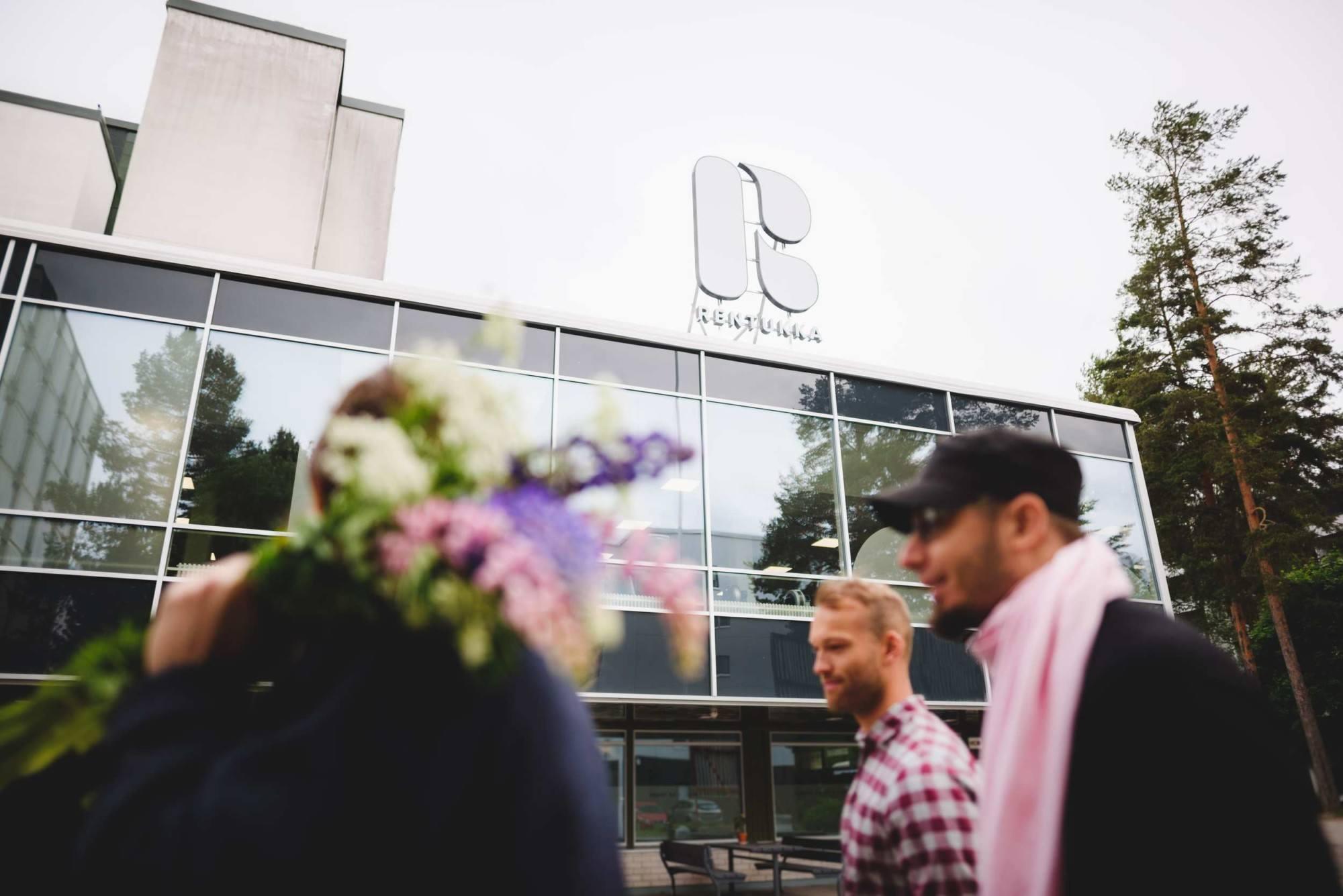 Yhteisötalo Rentukka. Kuvassa kolme henkilöä kävelemässä Rentukan edustalla, yhdellä heistä kimppu lupiineja olalla. Taustalla Rentukka-rakennus, jonka katolla suuri R-logo.