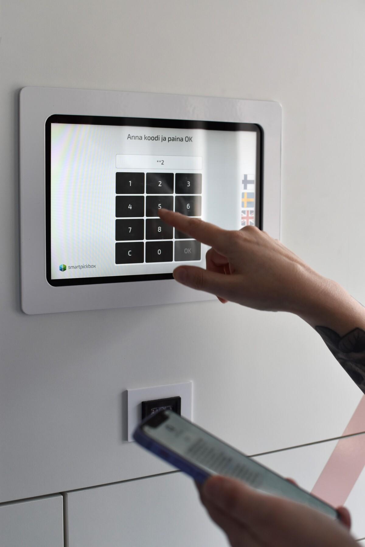 Kuvassa noutoautomaatin kosketusnäyttö, sormi näppäilee numeroa näytölle ja etualalla puhelin toisessa kädessä.