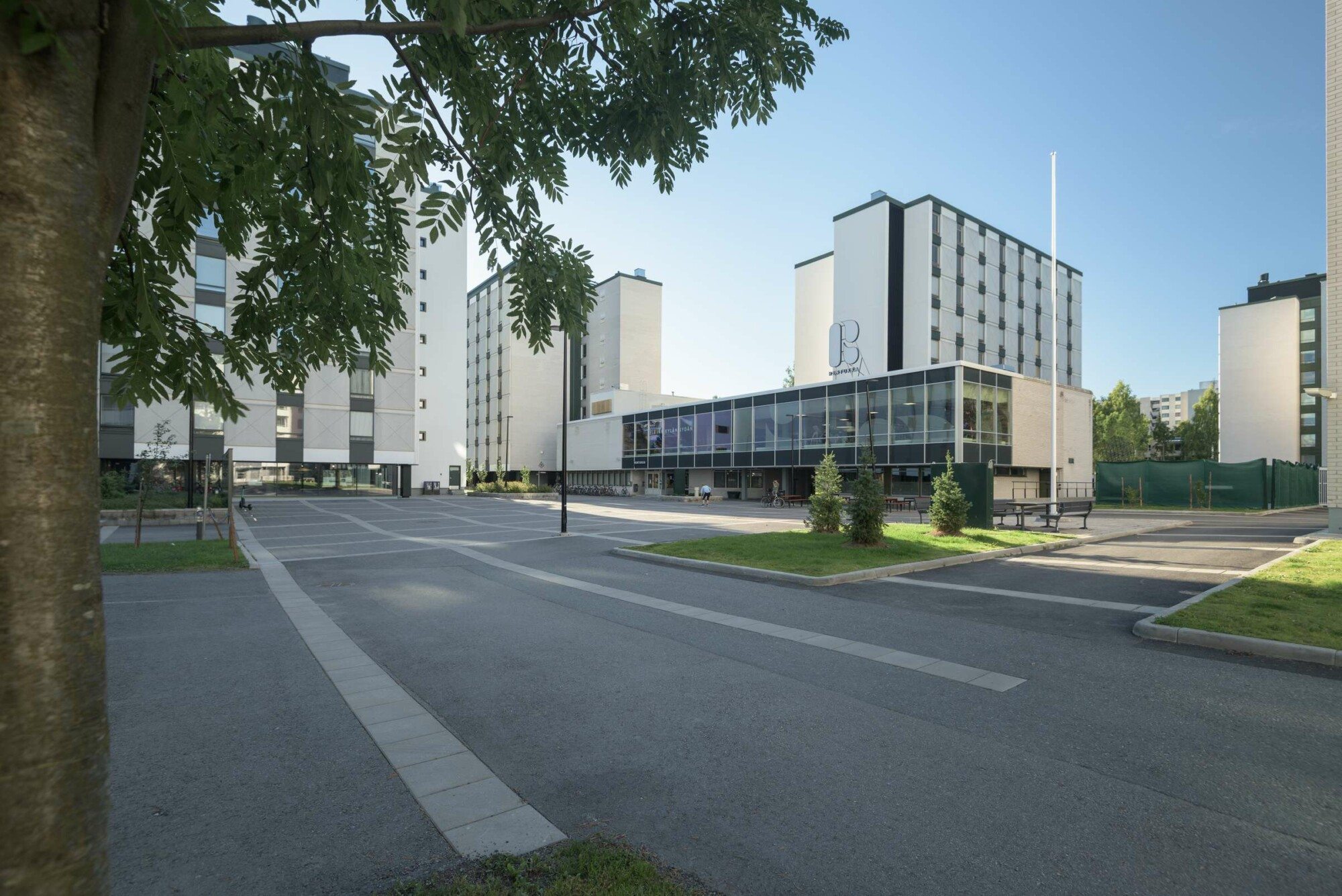 Aukio, jonka ympärillä korkeita, vaaleita kerrostaloja sekä matala rakennus, jonka katolla suuri R-logo. Aukion vierellä matalakasvuisia puita ja edustalla yksi puu.