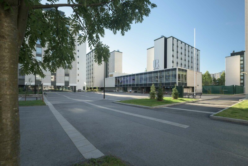 Aukio, jonka ympärillä korkeita, vaaleita kerrostaloja sekä matala rakennus, jonka katolla suuri R-logo. Aukion vierellä matalakasvuisia puita ja edustalla yksi puu.