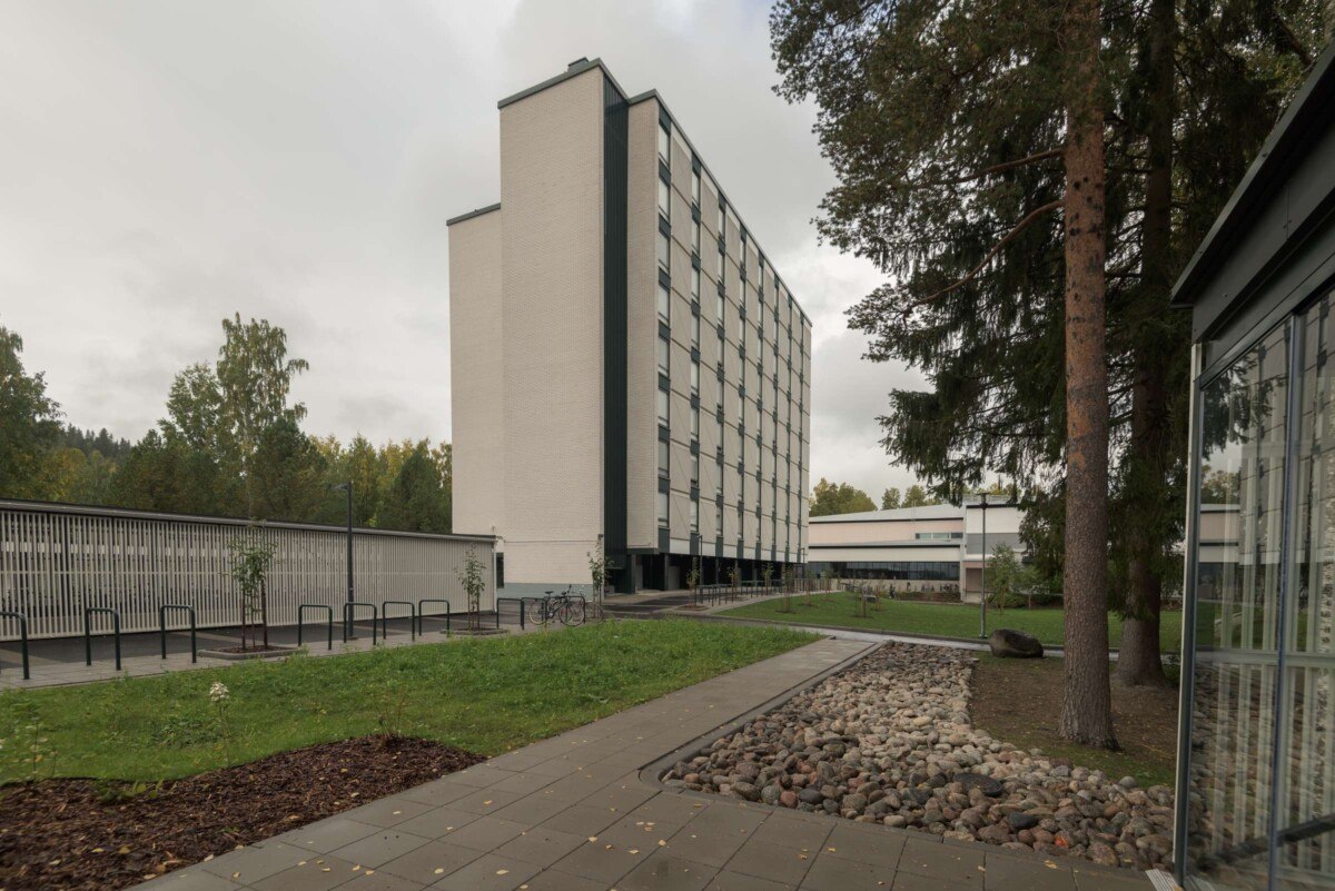 The nine-storey E-building in the Kortepohja Student Village. Kortepohja library in the background. Tree plantings and paving in the front. Bicycle parking on the left side.