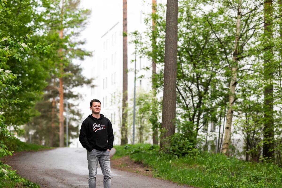 Henkilö seisoo metsäisellä kävelytiellä ja katsoo kameraan. Taustalla vaalea kerrostalo.