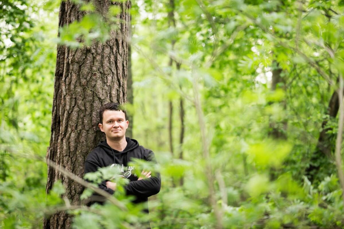 Henkilö nojaa puuta vasten ja katsoo hymyillen kameraan. Taustana vaalean vihreä metsä ja edustalla puun oksia.